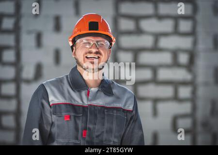 Porträt eines fröhlich lächelnden bärtigen kaukasischen Arbeiters in Overall, Schutzhelm und Schutzbrille auf einer Baustelle Stockfoto
