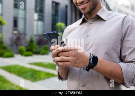 Nahaufnahme der Hände des Mannes, Geschäftsmann im Hemd, der das Telefon hält, App auf dem Smartphone nutzt, in der Stadt vor dem Bürogebäude spazieren geht. Stockfoto