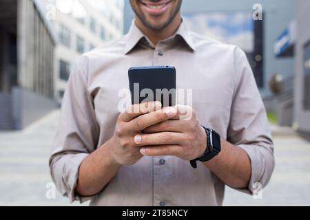 Nahaufnahme der Hände des Mannes, Geschäftsmann im Hemd, der das Telefon hält, App auf dem Smartphone nutzt, in der Stadt vor dem Bürogebäude spazieren geht. Stockfoto