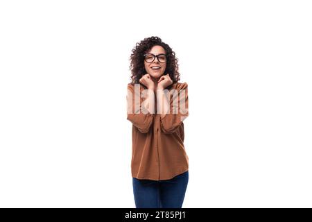 Die junge angenehm überraschte, fröhliche, schlanke Frau mit lockigem Haarschnitt trägt ein braunes Hemd Stockfoto