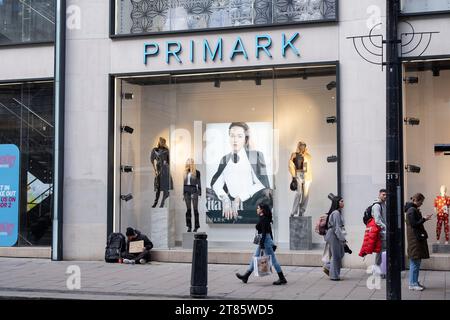 Einkäufer und Besucher vor der Tür gehen an einem Mann vorbei, der auf dem Bürgersteig sitzt und um Geld bittet, um Geld für ein Hostel außerhalb der Bekleidungsmarke Primark in der Oxford Street zu bezahlen, am 13. November 2023 in London, Großbritannien. Die Oxford Street ist ein bedeutendes Einkaufszentrum im West End der Hauptstadt und ist Europas geschäftigste Einkaufsstraße mit rund einer halben Million Besuchern der rund 300 Geschäfte, von denen die meisten Mode- und High Street-Bekleidungsgeschäfte sind. Stockfoto