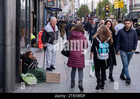 Als Käufer und Besucher auf der Oxford Street vorbeikommen, sitzt am 13. November 2023 in London, Großbritannien, eine Frau auf dem Bürgersteig und bettelt um Geld. Die Szene veranschaulicht die soziale Diskrepanz im Vereinigten Königreich mit einigen Menschen, die im Vergleich zu anderen in relativem Wohlstand leben. Stockfoto