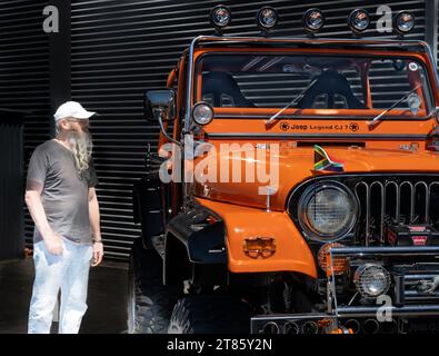 Mount Maunganui Neuseeland - 12. November 2023; Mann mit langem grauen Bart, der auf das hellorange Geländefahrzeug Jeep Legend CJ 7 blickt Stockfoto