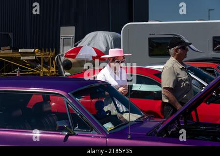 Mount Maunganui Neuseeland - 12. November 2023; All USA Car Day - Frau mit herzförmiger Sonnenbrille und rosafarbenem Hut spaziert zwischen Autos. Stockfoto