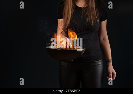 Brünette Frau mit tätowierten Händen, in Leder-Leggings und T-Shirt. Halten Sie eine Wok-Pfanne mit Feuer vor schwarzem Hintergrund. Kochkonzept. Nahaufnahme Stockfoto