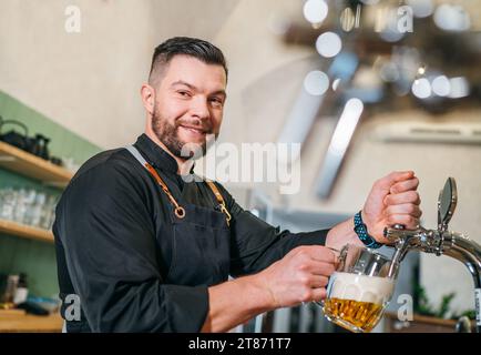 Lächelnder, stilvoller Bärenbarmann in Uniform mit Schürze, der in die Kamera blickt, während er an der Bar den Becher mit frischem Lagerbier anklopfte. Erfolgreiche Leute, Stockfoto