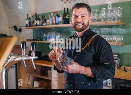 Porträt eines fröhlich lächelnden bärtigen Barkepers in schwarzer Uniform mit Schürze, der das Bierglas an der Bar abwischt. Erfolgreiche Mitarbeiter, harte Arbeit, Stockfoto