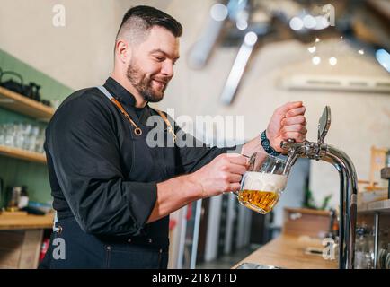 Ein lächelnder, stilvoller Bärenbarmann in schwarzer Uniform mit einer Schürze, der frisches Lagerbier in einen Glasbecher an der Bar zapft. Erfolgreiche Leute, Bierkonzern Stockfoto