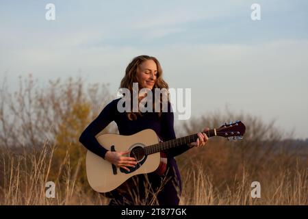 Schöne junge, lächelnde Frau, die eine Akustikgitarre spielt, während sie im Feld zwischen hohem, getrocknetem Gras steht. Talentierter Musiker. Sonnenuntergang im Herbst. Romantische Mo Stockfoto