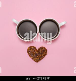 Kaffee kreative Liebe Emoji. Lächeln Sie von zwei Tassen schwarzen Espresso und gesunden rohen herzförmigen Keks als Mund. Lächelndes Gesicht mit Augen auf Rosa Stockfoto