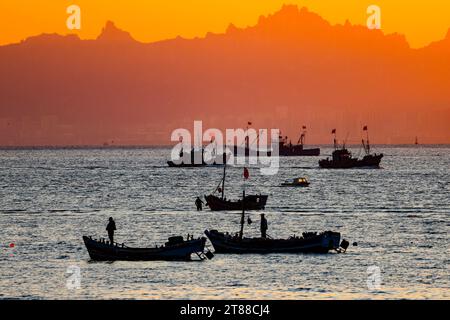 QINGDAO, CHINA - 18. NOVEMBER 2023 - Fischerboote segeln bei Sonnenuntergang an der Küste der Insel Gujia im New Areal der Westküste von Qingdao, Shandong Prov Stockfoto
