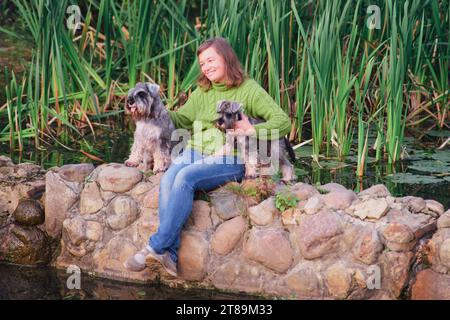 Niedliche Frau mit zwei kleinen Schnauzer-Hunden, die auf einem Felsen in der Nähe des Sees sitzen Stockfoto