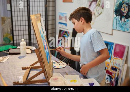 Hübscher Schuljunge, der ein Bild auf Leinwand zeichnet, steht neben einer hölzernen Staffelei in einer kreativen Kunstwerkstatt. Talentiertes Kind, inspirierter Künstler Maler, der seine genießt Stockfoto