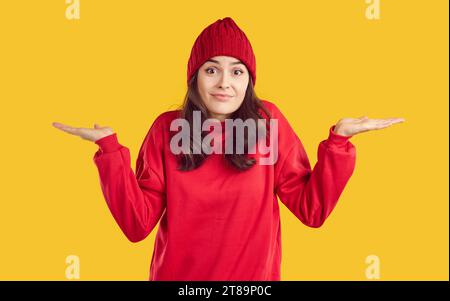 Ahnungslose Frau in rotem Sweatshirt und Hut zuckt Schultern isoliert auf gelbem Hintergrund Stockfoto