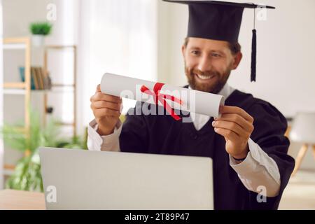 Glücklicher Student, der den Online-Abschluss abgeschlossen hat, sitzt am Laptop und hält das Diplom in der Hand Stockfoto