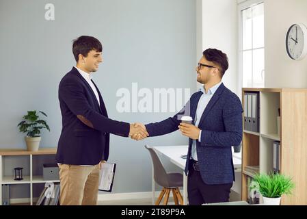 Zwei glückliche junge Geschäftsleute stehen im Büro, lächeln und schütteln die Hände Stockfoto