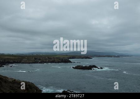 Felsige Küste mit rauem Wasser unter einem bewölkten Himmel, mit einer hügeligen Landschaft im Hintergrund Stockfoto