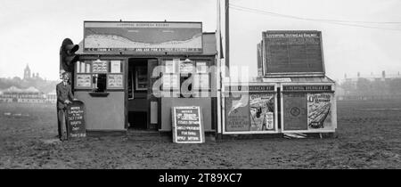 Ein Buchungsbüro der Liverpool Overhead Railway in einer Hütte auf einer schlammigen Aintree Racecourse, Liverpool, Merseyside, England, Großbritannien, um 1930. Auf Plakaten und Hinweistafeln wird für die Fahrkarten und Genehmigungen der Eisenbahn geworben – insbesondere um die Ozeanliner an den Docks zu sehen. Die RMS Scythia, die RMS Adriatic und die SS Doric werden von einem Pappausschnitt einer Eisenbahnwache (links) gehalten. Ein Zweig der Freibahn wurde 1906 eröffnet und diente einige Jahre lang der Rennbahn. Die Liverpool Overhead Railway war die erste elektrische Hochbahn der Welt. Die Bahnstrecke wurde 1956 geschlossen. Stockfoto