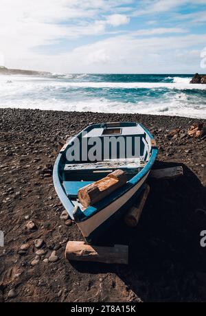 Landschaftsfoto des blauen alten Bootes (Schiff) auf vulkanischem schwarzem Sandstrand mit schönen Wellen im Ozean auf Hintergrund - Lanzarote Island. Landschaftsaufnahme Stockfoto