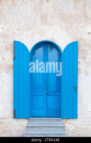 Echt blaue Tür mit blauen Fensterläden an der weißen Wand mit Rissen und abblätternder Farbe in Santa Maria di Leuca, Salento, Provinz Lecce, Apulien, Italien Stockfoto