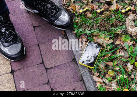 Smartphone mit kaputtem Display liegt zu den Füßen einer Person auf dem Asphalt Stockfoto