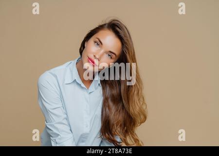 Schwüle Frau mit voluminösem Haar in blauer Bluse, die den Kopf auf beigem Hintergrund neigt Stockfoto