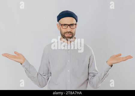 Ein bärtiger Mann in Brille, blauer Mütze und grauem Hemd steht vor einem weißen Hintergrund und drückt Verwirrung mit ausgestreckten Händen aus. Stockfoto