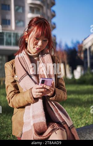 Junge lächelnde elegante asiatische Frau, die Schal in der Großstadt mit Handy-Plattform-Anwendungen trägt. Lächelnde Frau, die ihr Smartphone im Freien hält Stockfoto