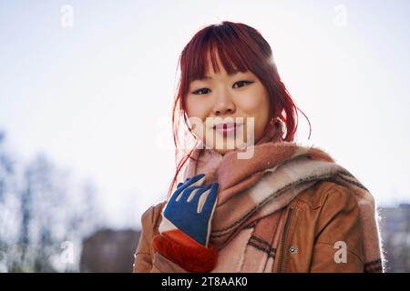 Junge selbstbewusste lächelnde asiatische Frau, die auf der Straße steht, Porträt. Stolze Frau, die einen Schal trägt, posiert in der Großstadt draußen in die Kamera. Stockfoto