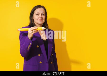 Eine Frau mittleren Alters, die einen violetten Blazer trägt, hält einen riesigen Bleistift auf gelbem Hintergrund Stockfoto