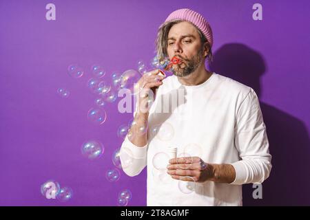 Sorgloser Mann mittleren Alters, der Seifenblasen vor violettem Hintergrund bläst Stockfoto