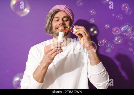 Sorgloser Mann mittleren Alters, der Seifenblasen vor violettem Hintergrund bläst Stockfoto