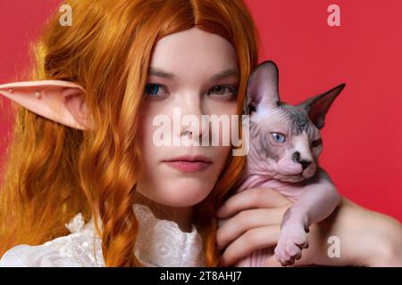 Nahaufnahme einer ernsten jungen Frau Cosplay-Elfe mit lockig rotem Haar. Elf in weißem Sommerkleid umarmt Sphinx-Kätzchen und blickt in die Kamera Stockfoto