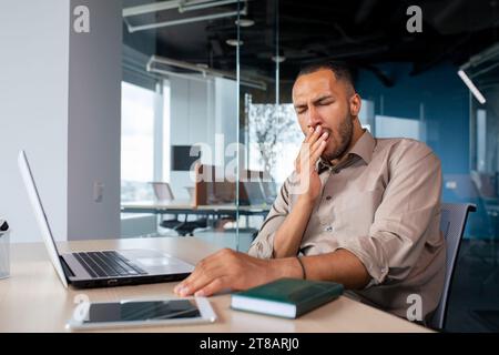 Gelangweilter arabischer Geschäftsmann, der am Tisch vor dem Laptop im Büro sitzt, gähnt, seinen Mund mit der Hand bedeckt, überarbeitet, Schlafmangel, Burnout. Stockfoto