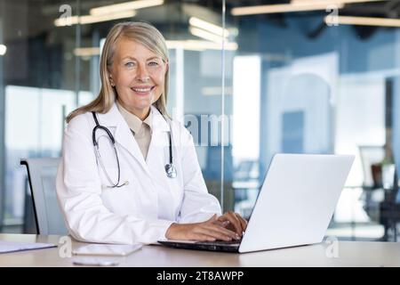 Porträt der Reifen erfahrenen Ärztin, grauhaarige ältere Frau im weißen medizinischen Mantel lächelnd in die Kamera, konsultierte Patienten aus der Ferne, sitzend am Tisch mit Laptop in der Klinik. Stockfoto