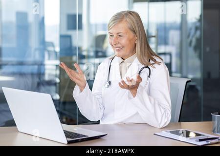 Reife erfahrene Ärztin spricht fernab mit Videoanruf, grauhaarige Krankenhausarbeiterin berät Patienten, die im modernen Klinikbüro in weißem medizinischen Mantel sitzen. Stockfoto