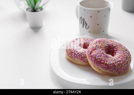 Donuts mit rosa Zuckerguss und farbigen Streuseln auf einem weißen Teller liegen auf einem weißen Tisch neben einer weißen Tasse mit schwarzen Punkten. Hochwertiges Foto auf dem l Stockfoto