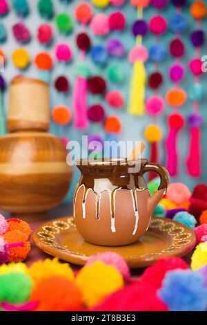 Mexikanische Champurrado heiße Schokolade in traditioneller rustikaler Jarritos-Becher mit Zimtstab und bunter Pompons-Girlande Stockfoto