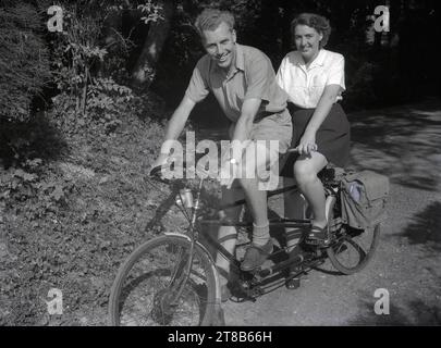 1950er Jahre, Geschichte, Radurlaub, Sommer und draußen auf einem Landweg, ein lächelndes Paar, beide in Shorts, sitzend auf einem Tandemfahrrad, Kent, England, Großbritannien. Auf dem Rücken sind die Leinwandtaschen der Ära. Stockfoto