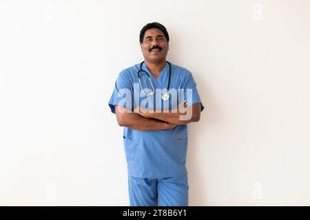 Medizinischer Mitarbeiter. Lächelnder Indischer Männlicher Arzt In Uniform Stehend Mit Gefalteten Armen Stockfoto
