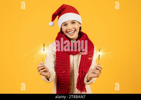 Eine junge Frau im Weihnachtsmann-Hut hält leuchtende Glitzer, gelber Hintergrund Stockfoto