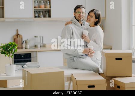 Liebevolles Paar, das einen Moment mit Kaffee in einer neuen Küche, umgeben von Umzugskartons, teilt Stockfoto