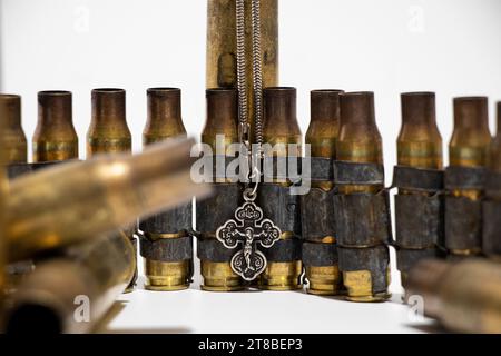 Gebrauchte Maschinengewehrgürtel und Patronen und ein silbernes Kirchenkreuz hängen an Kugeln auf weißem Hintergrund, Krieg und glaube an Gott Stockfoto