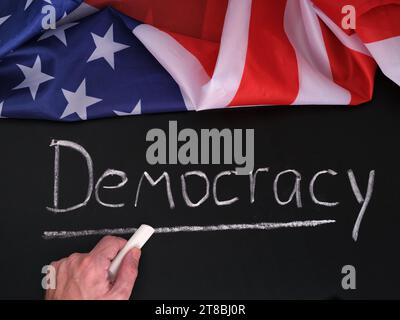 Die Flagge der USA, die auf einer Tafel liegt und das Wort Demokratie von einem Mann mit weißer Kreide geschrieben wird. Nahaufnahme Stockfoto