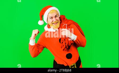 Glücklicher Weihnachtsmann mit einer Tasche voller Geschenke. Ein aufgeregter bärtiger Mann im Weihnachtsmannkostüm, der große Weihnachtssack mit Geschenken trägt. Lächelnder Mann mit Weihnachtsmütze Stockfoto