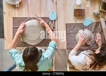 Blick von oben auf zwei Frauen, die mit Ton arbeiten, während sie in einem Töpferkurs handwerkliche Stücke herstellen. Bastel- und Hobby-Konzept. Stockfoto