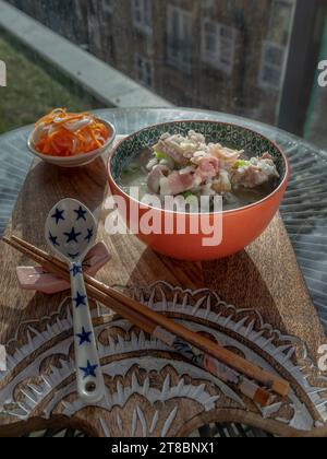 Heißer gekochter Reis oder Reisbrei mit Schweinerippchen Suppe und Schinken serviert mit eingelegten Karotten und Rettich auf hölzernem Schneidebrett. Gesundes Frühstück Stockfoto