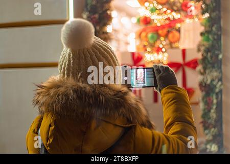 Eine Touristin mit Strickmütze fotografiert das festliche Neujahrsdekor auf ihrem Smartphone. Weihnachtsmarkt, Nachtbeleuchtung. Blick von hinten. Stockfoto