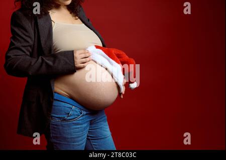 Porträt des großen Bauches einer graviden Frau, werdende Mutter, die Santa Hut auf ihren schwangeren Bauch legt, isoliert auf rotem Atelierhintergrund mit Stockfoto