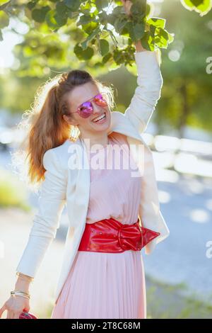 Lächelnde, stilvolle Frau in rosa Kleid und weißer Jacke in der Stadt mit Sonnenbrille. Stockfoto